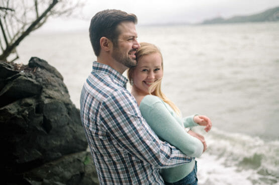 A man in a plaid shirt hugs a smiling woman in a light blue sweater as they stand by rocks near a cloudy, windswept shoreline. by Justin Salem Meyer