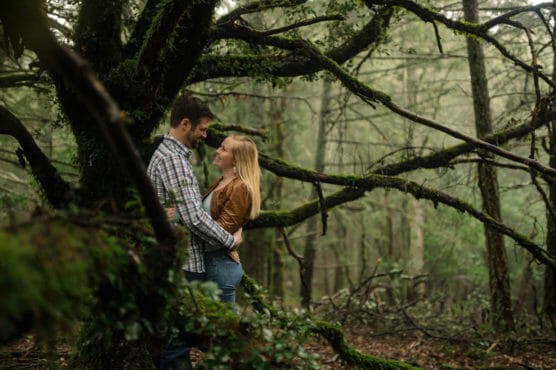 A man and woman stand close together, smiling and gazing into each other’s eyes beneath a large, mossy tree in a misty, green forest. by Justin Salem Meyer