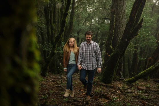 A couple walks hand in hand through a dense, green forest. The woman wears a brown jacket and jeans, and the man wears a plaid shirt and jeans. Tall trees with moss surround them, and the ground is covered in leaves. by Justin Salem Meyer