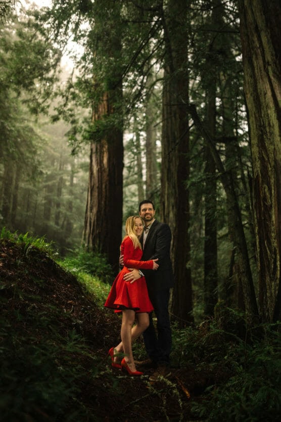 A couple stands together in a lush, misty forest. The woman wears a bright red dress and heels, while the man is dressed in a dark suit. They smile and embrace among tall trees and green foliage. by Justin Salem Meyer