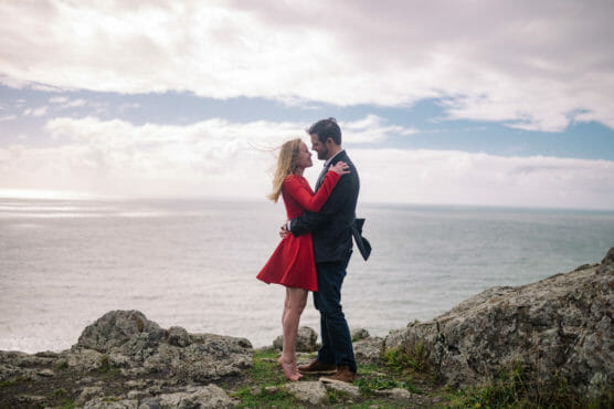A couple stands barefoot on rocky ground by the ocean, embracing and smiling at each other. The woman wears a red dress, and the man wears a dark suit. The sky is cloudy with patches of blue. by Justin Salem Meyer