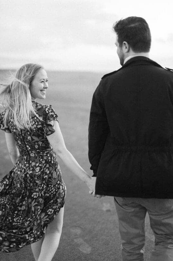 A smiling woman in a floral dress holds hands with a man in a dark jacket as they walk together on a beach. The black and white photo captures a joyful, candid moment. by Justin Salem Meyer