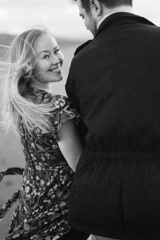 A woman with blonde hair and a floral dress smiles at a man beside her, who is turned away from the camera. The image is in black and white, capturing a joyful, candid moment outdoors. by Justin Salem Meyer