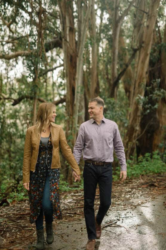 A couple walks hand-in-hand on a forest path, smiling at each other. The woman wears a brown jacket, floral dress, and boots, while the man wears a light purple shirt and dark jeans. Tall trees and greenery surround them. by Justin Salem Meyer
