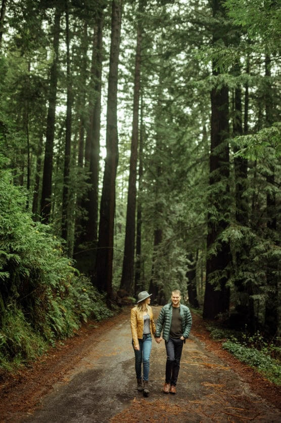 A couple walks hand in hand down a dirt path surrounded by tall, lush green trees in a forest. The ground is damp, and they are both dressed in jackets and hats, enjoying a peaceful moment in nature. by Justin Salem Meyer