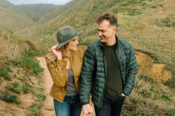 A smiling couple walks hand in hand along a dirt trail through green hills. The woman wears a brown jacket and gray hat; the man wears a dark green jacket. They look at each other affectionately. by Justin Salem Meyer