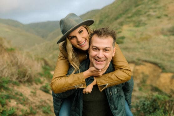 A smiling woman wearing a hat and brown jacket gives a man a piggyback ride outdoors, both looking at the camera with hills and greenery in the background. by Justin Salem Meyer