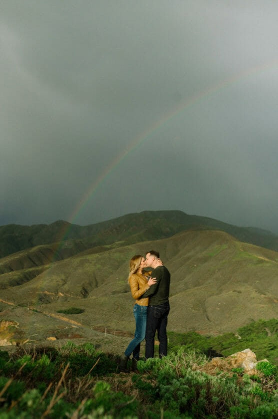 A couple embraces and kisses on a grassy hill with green mountains and a rainbow in the background under a cloudy sky. by Justin Salem Meyer
