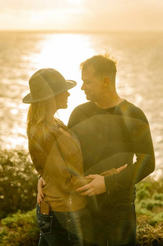 A couple stands close together outdoors at sunset, facing each other and smiling. The woman wears a hat and a brown jacket, and the man wears a dark long-sleeve shirt. Sunlight shines brightly over water in the background. by Justin Salem Meyer