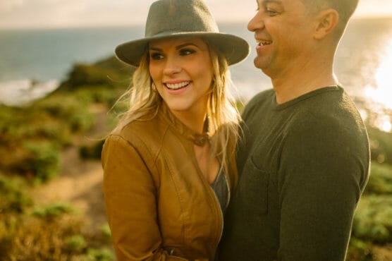 A smiling woman in a brown jacket and gray hat stands beside a smiling man in a dark sweater outdoors with greenery and the ocean in the background, lit by warm sunlight. by Justin Salem Meyer