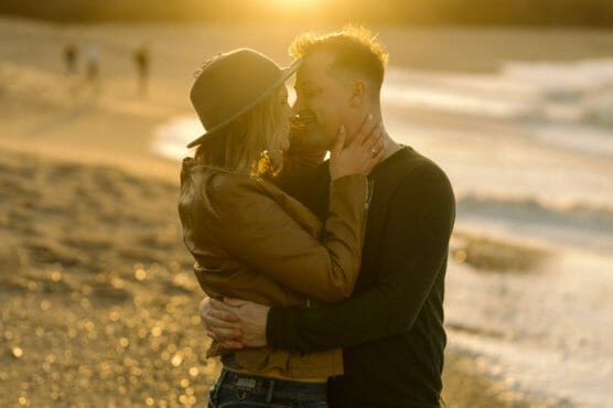 A couple embraces on a sandy beach at sunset, gazing into each other’s eyes and smiling. The warm, golden sunlight creates a romantic and dreamy atmosphere. by Justin Salem Meyer