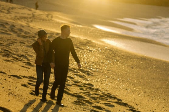 A couple walks hand in hand along a sandy beach at sunset, casting long shadows. The woman wears a hat and jacket, and the man wears a sweater. The ocean and waves are in the background with golden sunlight. by Justin Salem Meyer