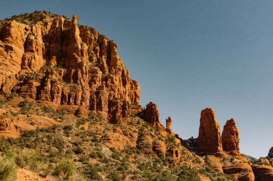Red rock formations rise against a clear blue sky in a desert landscape, with rugged cliffs and sparse vegetation dotting the rocky slopes. by Justin Salem Meyer