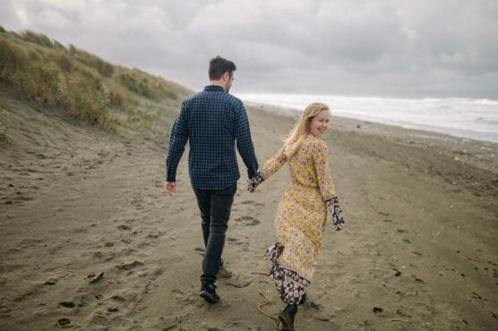 A couple holding hands walks along a sandy beach. The woman in a yellow floral dress looks back and smiles, while the man in a plaid shirt faces forward. Grassy dunes and a cloudy sky are in the background. by Justin Salem Meyer