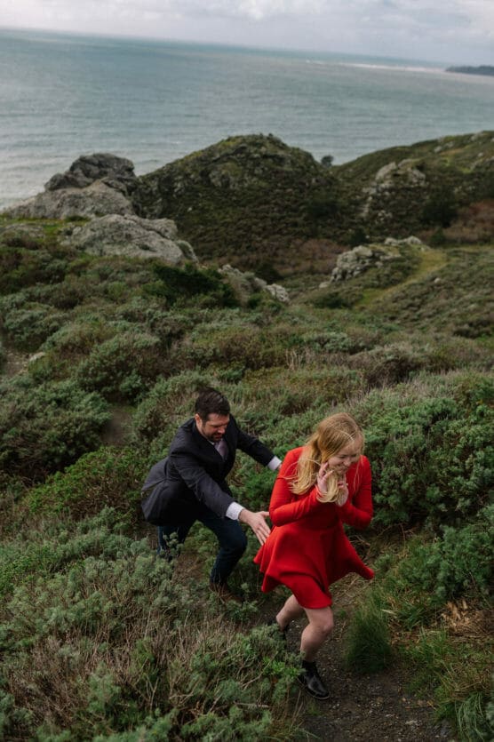 A man and woman walk playfully up a grassy, rocky hillside near the coast. The woman in a red dress leads, smiling, while the man follows closely behind her. The ocean and cloudy sky are visible in the background. by Justin Salem Meyer