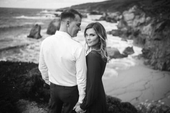 A couple stands hand in hand on a rocky coastline. The woman looks over her shoulder at the camera while the man gazes at her. The scene is in black and white, with waves crashing against the shore in the background. by Justin Salem Meyer