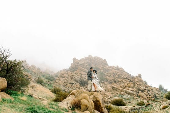 A couple stands close together on a rocky hilltop, wrapped in a blanket, surrounded by boulders and sparse greenery under a misty, overcast sky. by Justin Salem Meyer