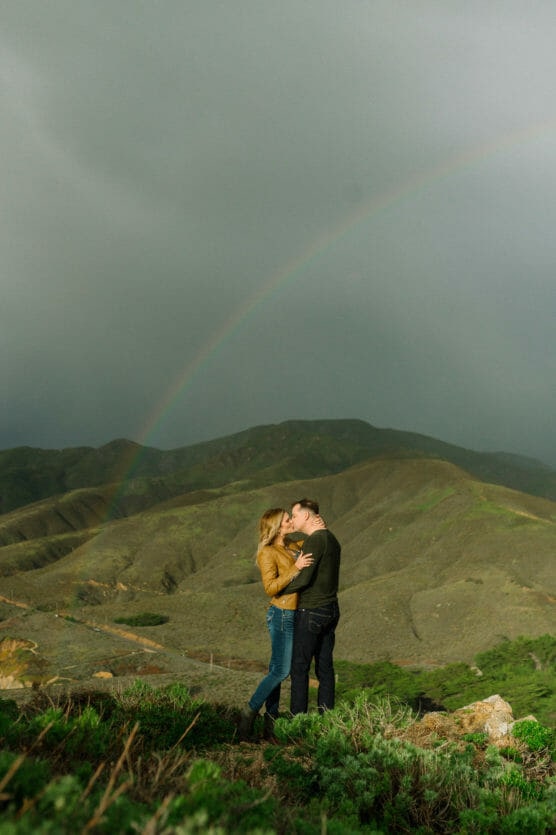 A couple embraces and kisses on a grassy hillside with green mountains in the background and a faint rainbow arching across a cloudy sky. by Justin Salem Meyer