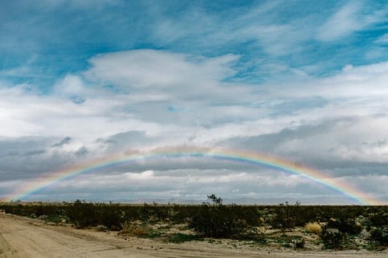 A vivid rainbow arcs across a cloudy blue sky above a flat, desert landscape with scattered shrubs and sandy dirt. by Justin Salem Meyer