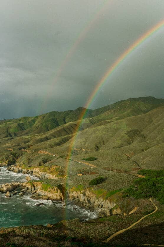 A vibrant double rainbow arcs over green hills along a rugged coastline, with waves crashing against rocky cliffs under a cloudy sky. by Justin Salem Meyer
