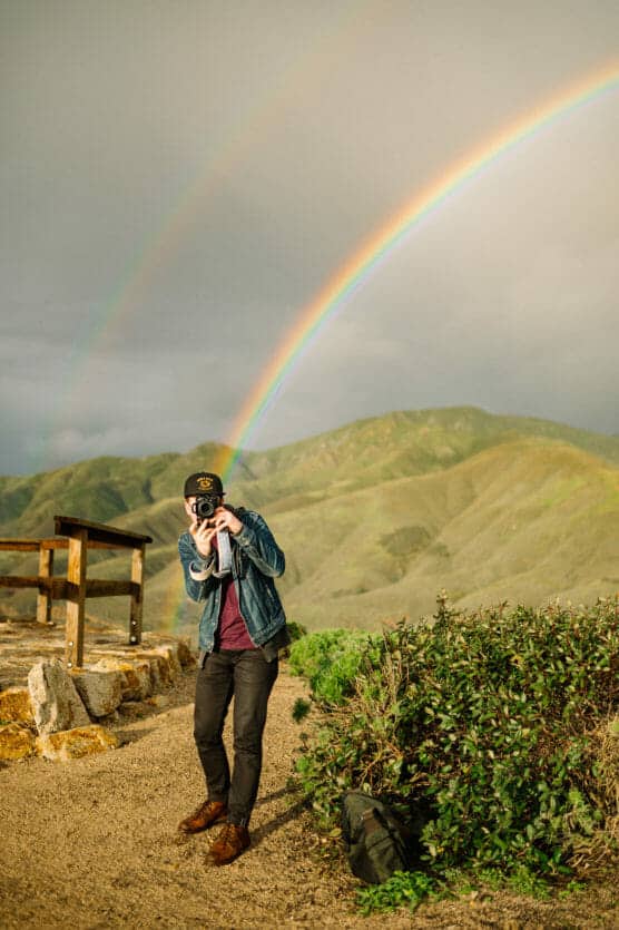 A person stands on a dirt path in front of green hills, holding a camera and smiling. Two vivid rainbows arc across the cloudy sky in the background. A backpack sits on the ground nearby. by Justin Salem Meyer
