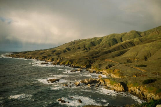 A rugged coastline with rocky cliffs and green hills beside the ocean under a cloudy sky, with waves crashing against the shore. by Justin Salem Meyer