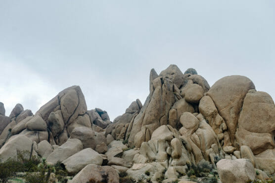 Large tan rock formations with rounded and jagged shapes rise against a cloudy sky, surrounded by sparse desert vegetation at the base. by Justin Salem Meyer