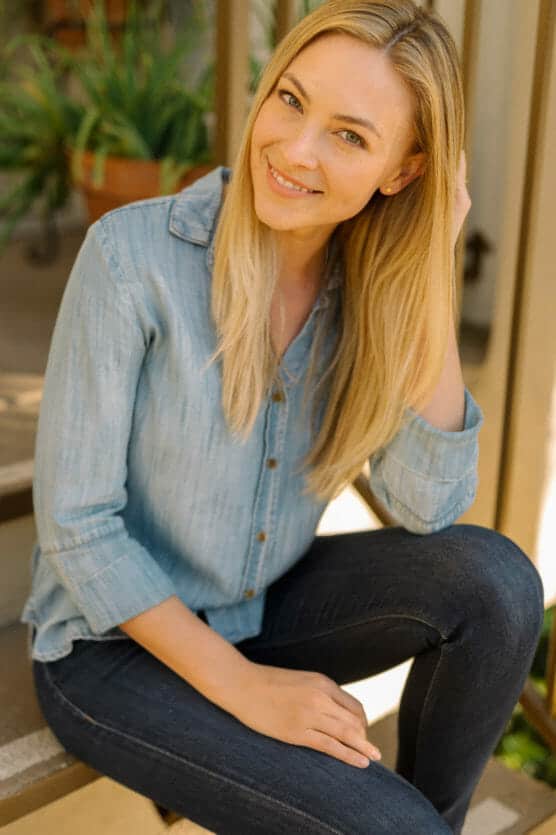 A woman with long blonde hair, wearing a light blue denim shirt and dark jeans, sits on outdoor steps, smiling at the camera and touching her hair. Green plants are visible in the background. by Justin Salem Meyer