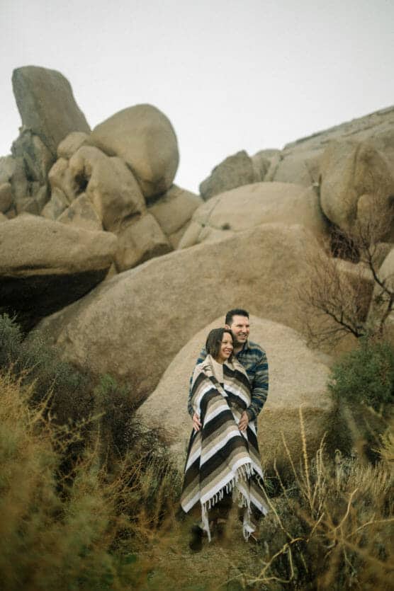 A couple stands close together outdoors, wrapped in a striped blanket, smiling in front of large, rugged boulders and sparse brush on a cloudy day. by Justin Salem Meyer