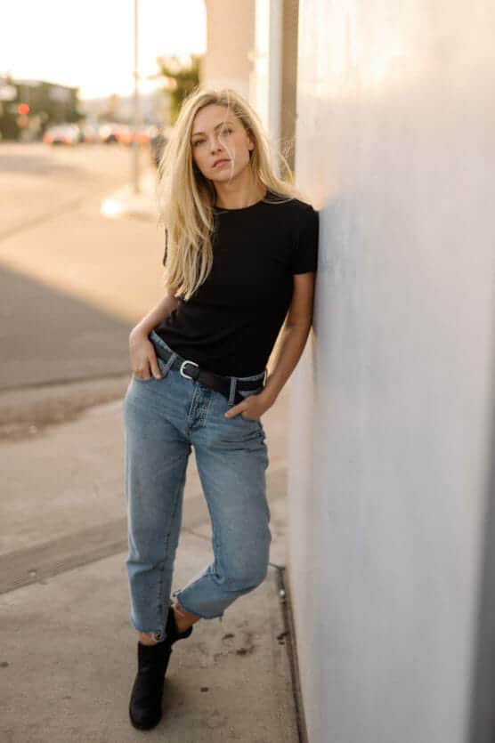 A woman with long blonde hair, wearing a black t-shirt, blue jeans, and black boots, leans casually against a light-colored wall on a city sidewalk with one hand in her pocket. by Justin Salem Meyer