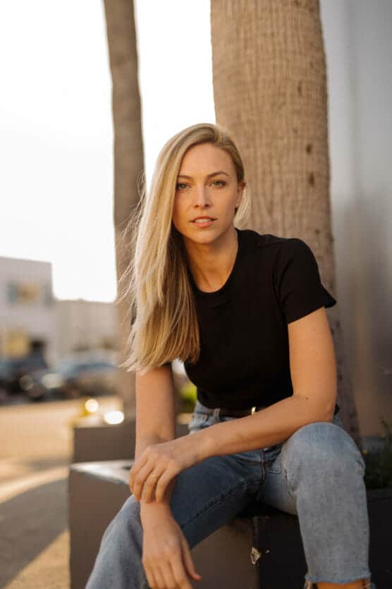 A woman with long blonde hair, wearing a black t-shirt and blue jeans, sits outdoors on a bench near a palm tree, looking at the camera in soft sunlight. by Justin Salem Meyer