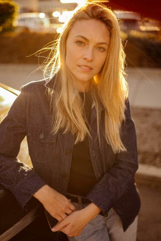 A woman with long blonde hair leans against a car outdoors, wearing a dark denim jacket and light jeans, looking at the camera. The photo is bathed in warm, golden sunlight. by Justin Salem Meyer