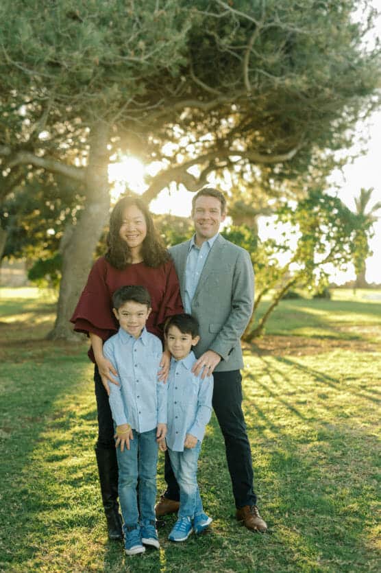 A family of four stands outdoors in a park with sunlight streaming through the trees. The parents stand behind their two young sons, all smiling at the camera on a sunny day. by Justin Salem Meyer