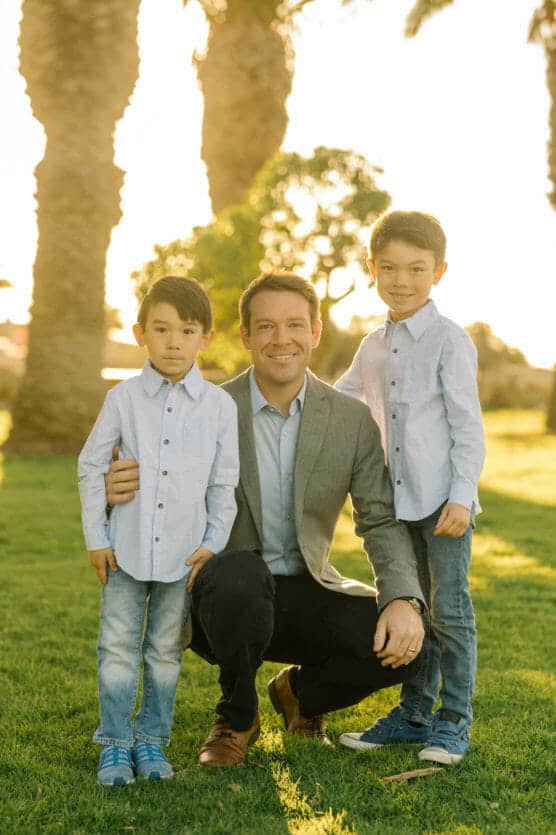 A man in a gray blazer kneels on grass, smiling with two young boys standing on either side. All wear light blue shirts and jeans. Palm trees and sunlight are in the background. by Justin Salem Meyer