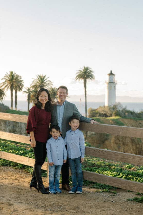 A family of four poses outdoors near a wooden fence, with palm trees and a white lighthouse in the background. The parents stand behind their two young sons, all smiling and dressed in casual, coordinated outfits. by Justin Salem Meyer