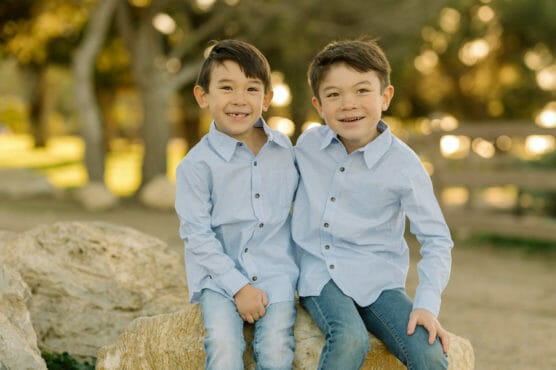 Two young boys with short brown hair, wearing matching light blue button-up shirts and jeans, sit close together on a large rock outdoors, smiling at the camera. Trees and sunlight are visible in the background. by Justin Salem Meyer