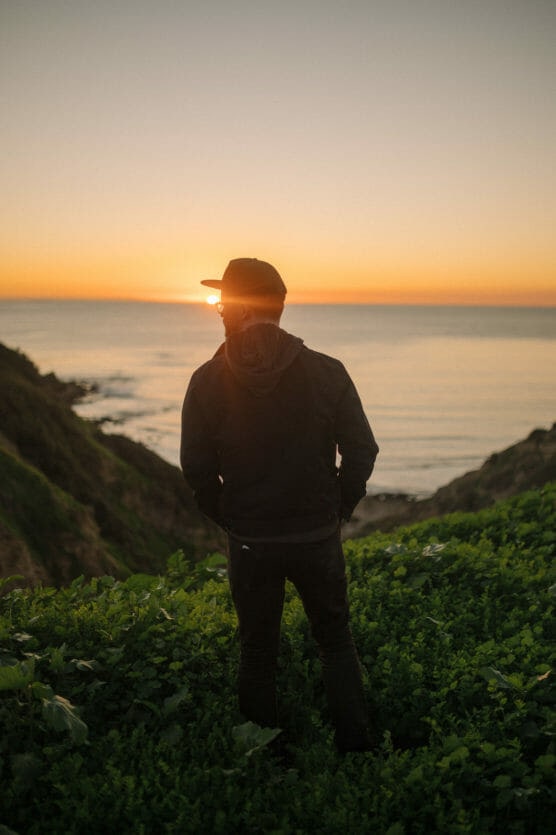 A person wearing a black jacket and cap stands on a grassy hill overlooking the ocean at sunset, with the sun low on the horizon and casting a warm orange glow across the sky and water. by Justin Salem Meyer
