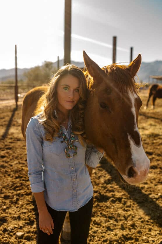 A woman with long wavy hair in a denim shirt stands next to a brown horse with a white blaze on its face, gently holding its head, in a sunlit outdoor paddock. by Justin Salem Meyer