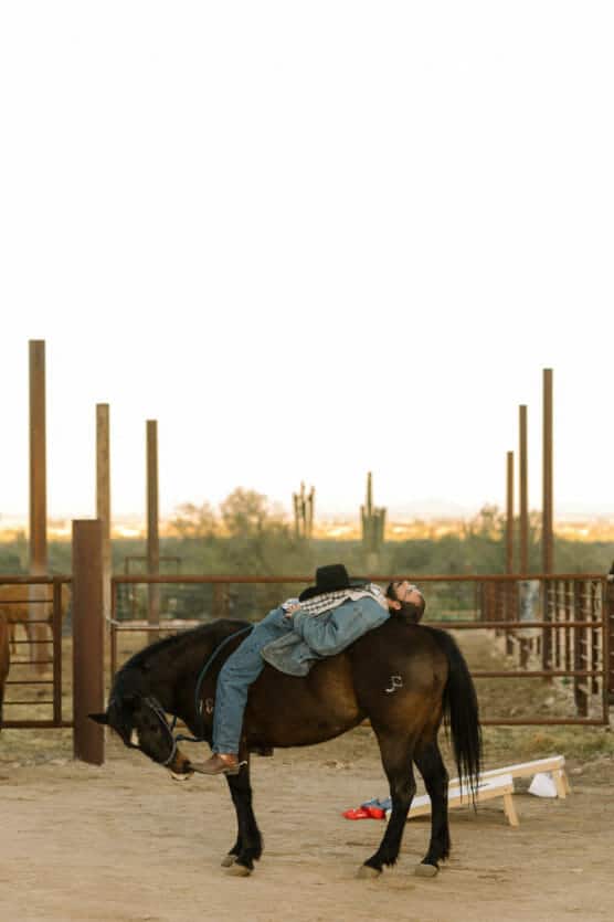 A person dressed in jeans, boots, and a cowboy hat lies on their back across a dark brown horse in a sandy corral, with desert plants and metal fencing in the background. by Justin Salem Meyer