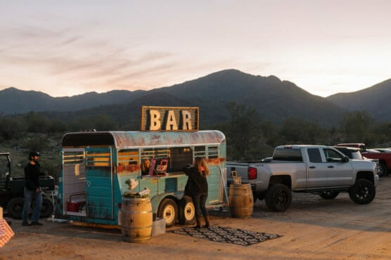 A teal trailer converted into a bar stands outdoors at sunset, with a large BAR sign on top. A person is serving drinks while a pickup truck is parked nearby. Mountains and a soft sky are in the background. by Justin Salem Meyer