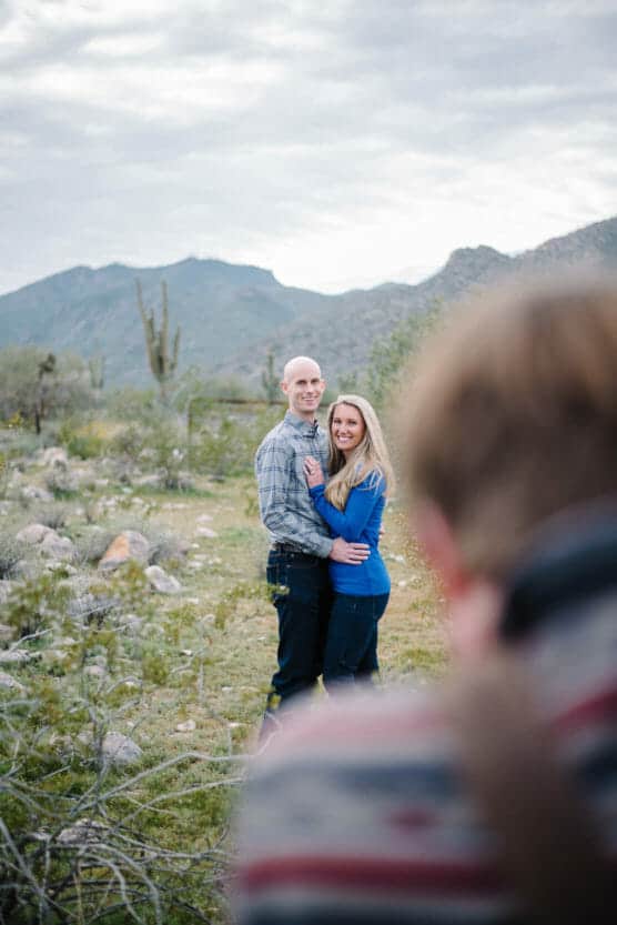 A couple poses together outdoors on a rocky path with desert plants and mountains in the background. The man holds the woman, both smiling at the camera. The blurred foreground shows part of a photographer capturing the moment. by Justin Salem Meyer