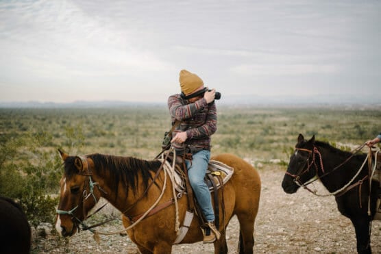A person in a yellow beanie and patterned jacket sits on a brown horse, taking a photo with a camera in a vast, open landscape, while another horse stands nearby. by Justin Salem Meyer