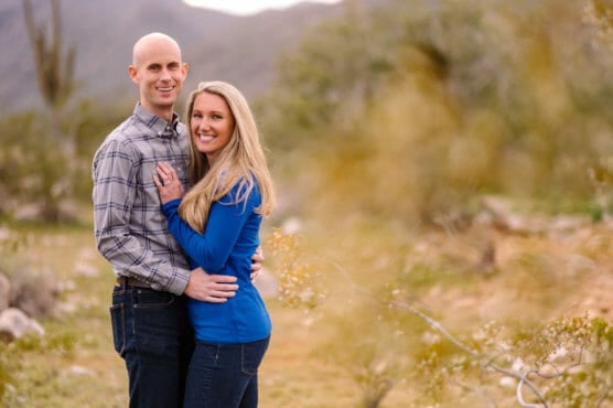 A smiling couple stands close together outdoors in a natural setting, with trees and mountains in the background. The woman wears a blue top and the man wears a plaid shirt. Both look relaxed and happy. by Justin Salem Meyer