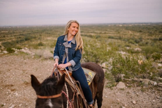 A woman with long blonde hair, wearing a denim jacket and jeans, smiles while sitting on a brown horse in a rocky, open landscape with greenery and a distant horizon under a cloudy sky. by Justin Salem Meyer