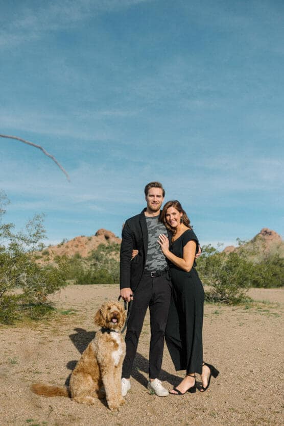 A smiling couple stands together in a desert landscape under a blue sky, with a happy brown and white dog sitting at their feet. Rocky hills and sparse green bushes are in the background. by Justin Salem Meyer