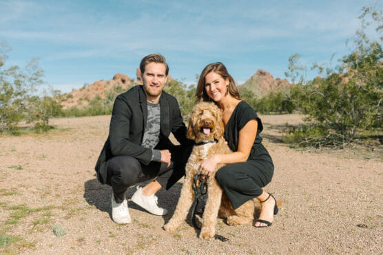 A man and woman crouch beside a fluffy brown dog, smiling outdoors on a sunny day. Rocky hills and scattered green bushes are visible in the background under a blue sky. by Justin Salem Meyer