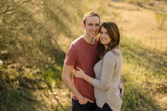 A young couple stands closely together outdoors, smiling at the camera. The man wears a red t-shirt and the woman wears a light gray sweater. Sunlight filters through trees in a grassy, natural setting. by Justin Salem Meyer