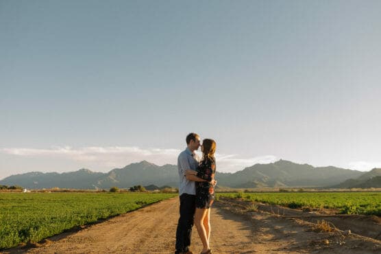 A couple stands embracing on a dirt road in a rural landscape, with green fields and distant mountains under a clear, blue sky at sunset. by Justin Salem Meyer