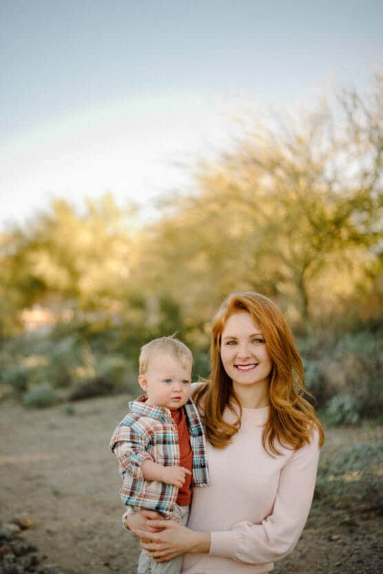 A woman with long red hair smiles while holding a toddler in her arms outdoors. The background features desert plants and trees under a clear, sunny sky with a faint rainbow. by Justin Salem Meyer