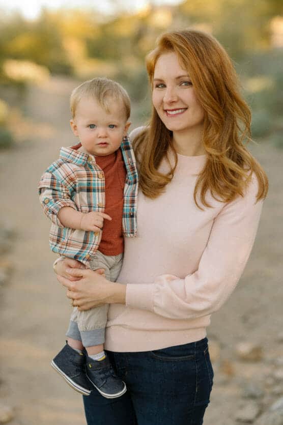 A smiling woman with long red hair, wearing a light pink sweater and dark jeans, holds a toddler boy in her arms. The boy wears a plaid shirt, rust-colored shirt, and gray pants. They are outdoors on a dirt path. by Justin Salem Meyer